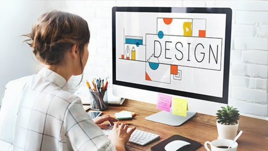 A lady creating.a design on a desktop with the monitor displaying the word "Design" and simple shapes around it.