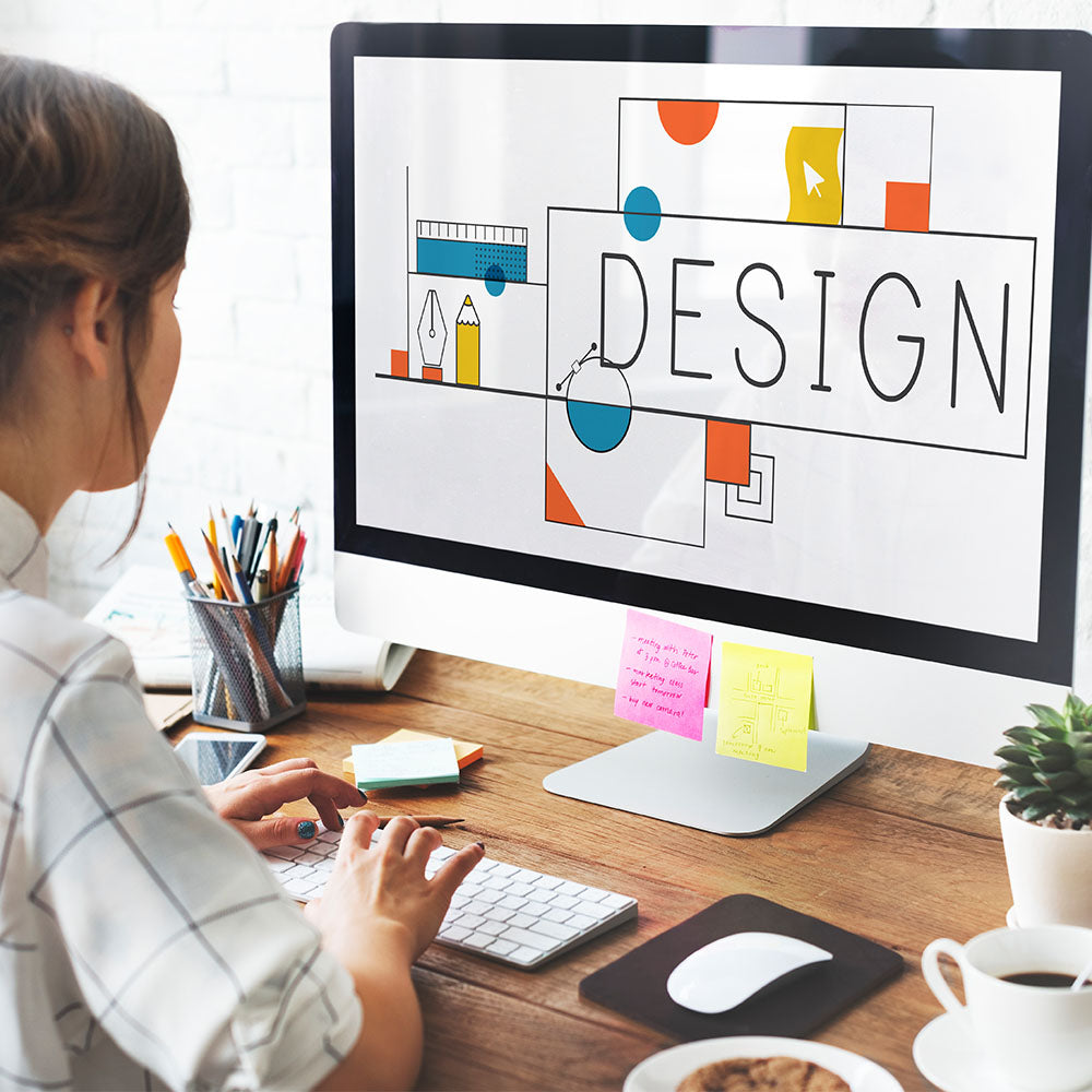 A person seated at a desk with a computer monitor displaying the word 'DESIGN' in stylized lettering, surrounded by colorful shapes and graphics.